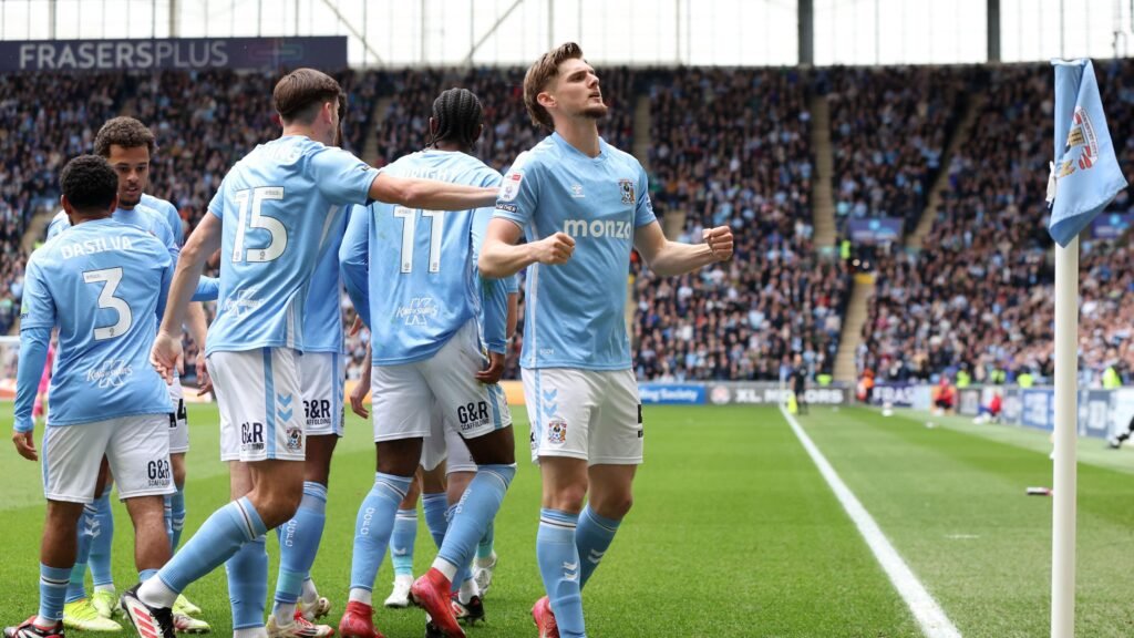 Coventry City players in sky-blue kits celebrate a goal near the corner flag in front of a packed stadium crowd during a football match.